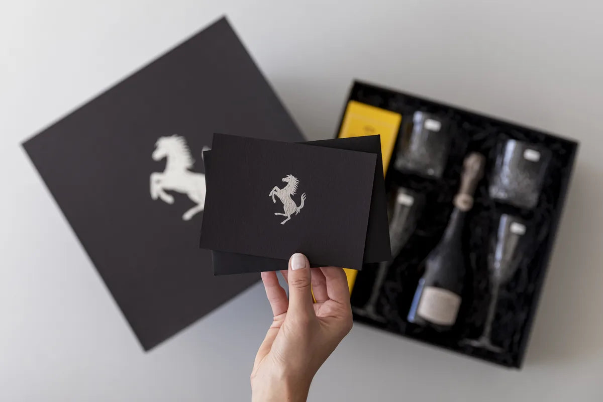 A Ferrari-branded black LÂPACH gift box and matching card, with champagne and tumblers set into fitted black filler.